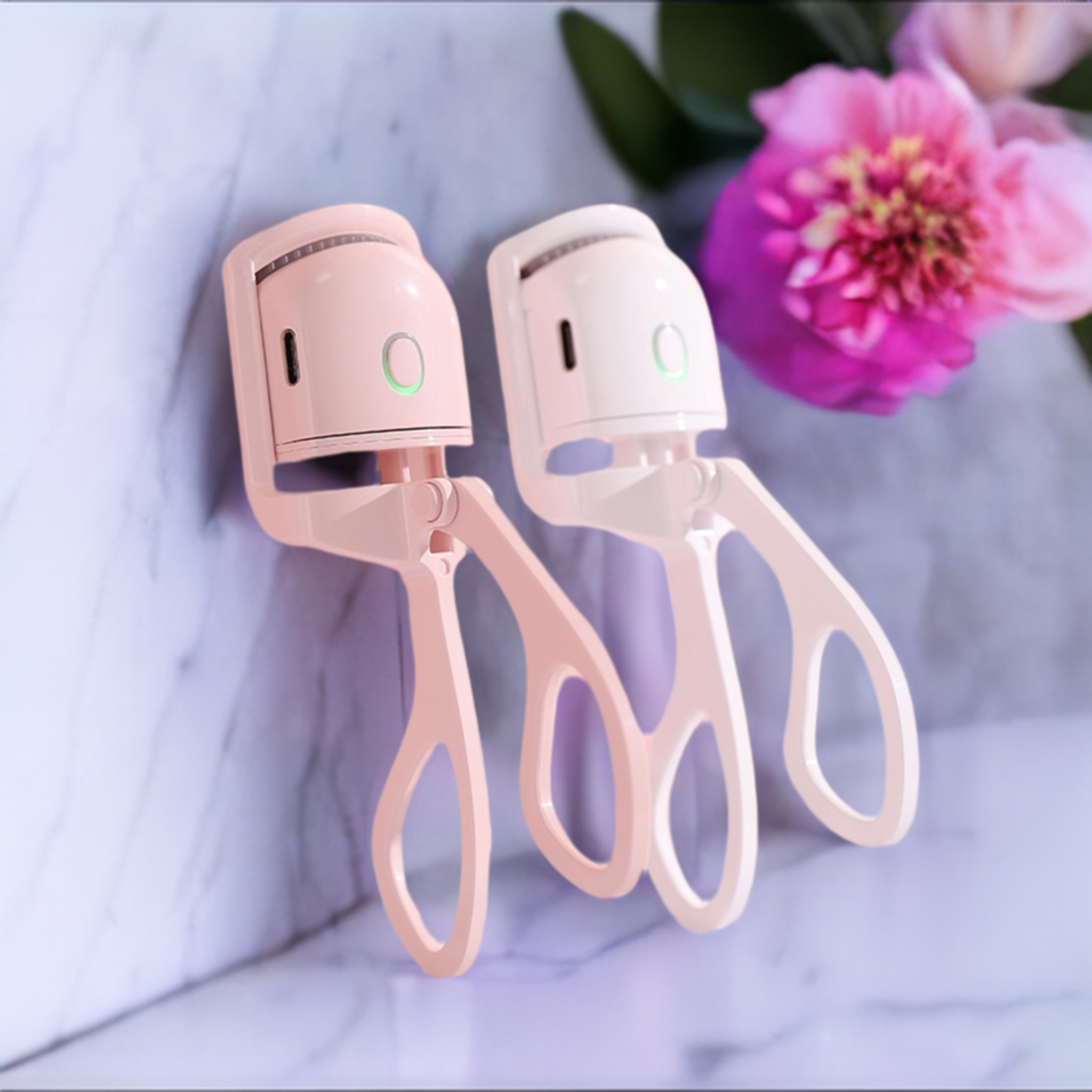 Pair of heated electric eyelash curlers on a marble table next to a flower.