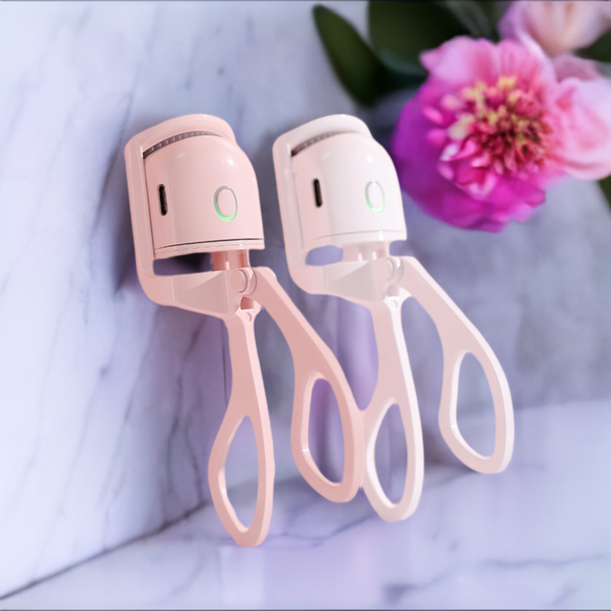 Pair of heated electric eyelash curlers on a marble table next to a flower.