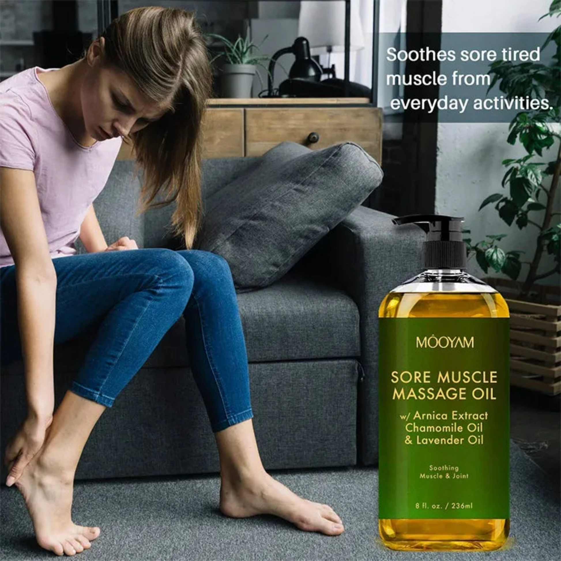 A woman sitting on a couch with a bottle of sore muscle natural massage oil rubbing her foot.