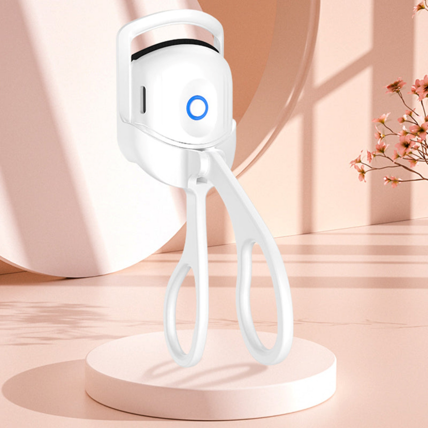 White electric heated eyelash curler standing upright on a peach-colored table next to a flower.